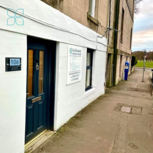 A small white shop with a black door has a white sign with black and blue writing on it
