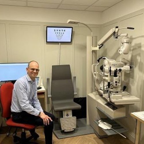 A smiling man sits on a red chair in an opticians office, complete with a grey chair and large eye testing equipment