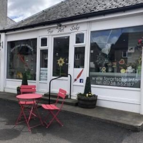 A small white building with large windows has a sign reading For Arts Sake written in cursive, and sits next to outdoor chairs