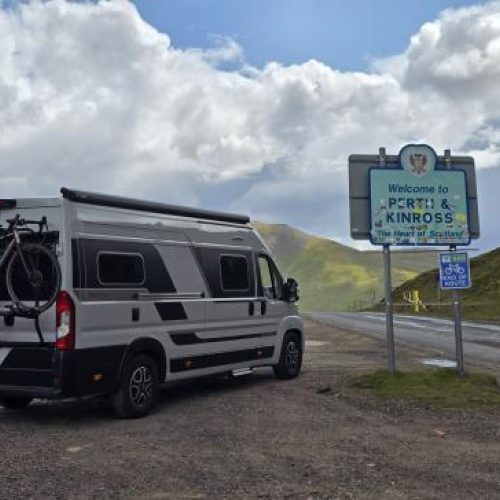 A large silver van with a bike attached to the back is parked next to a blue Perth and Kinross sign in a highland landscape