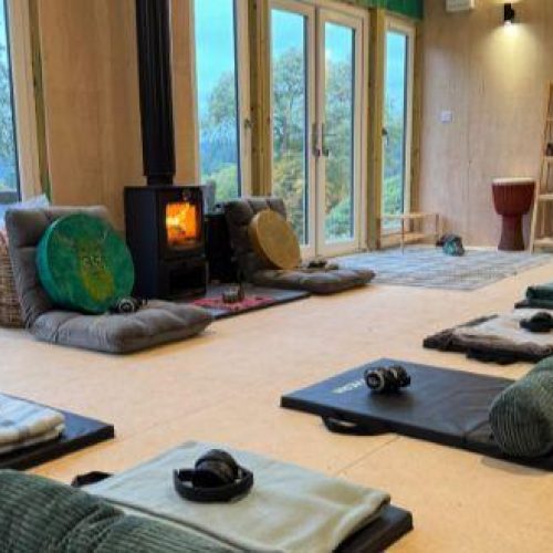 A light room with tall windows is filled with yoga mats, pillows, and headphones all lined up on the floor in front of a fireplace
