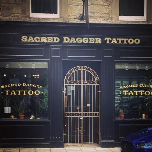 A black shop with an iron fence in front of the door between two large windows decorated with plants and gold lettering