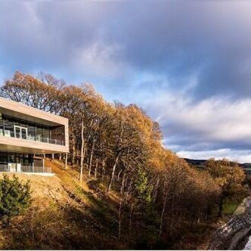 A square building with a glass panelled balcony its among autumnal trees on a hill overlooking a river