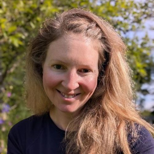 A blonde woman wearing a purple shirt smiles in front of a tree with green leaves