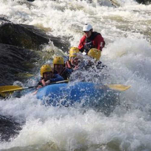 A group of people wearing wet suits and yellow helmets hold ores and sit in a raft going through large river rapids