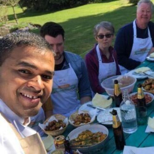 A group of people wearing matching white aprons sit around an outside bench covered in a spread of an array of food and wine