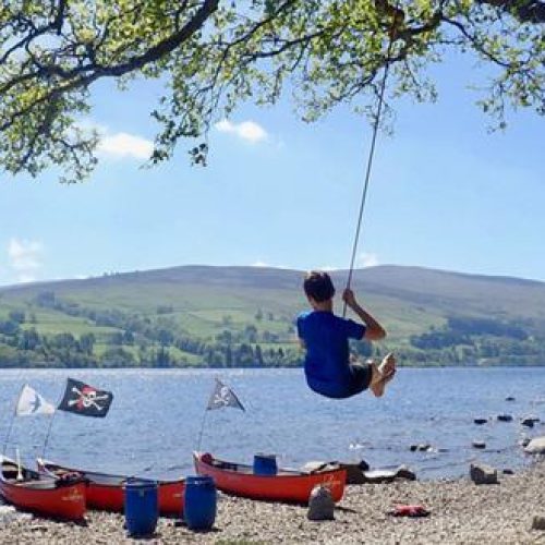 A man swings from a rope tied to a tree next to a loch with small red boats on the shoreline