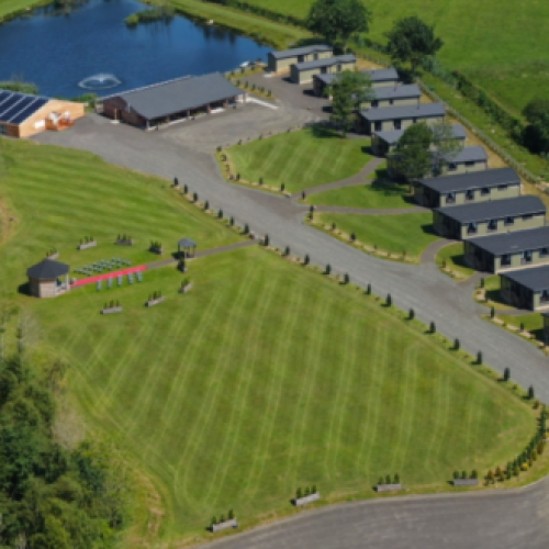 Several small green cabins are lined up next to a small pond and a large open lawn with a small hut in the centre
