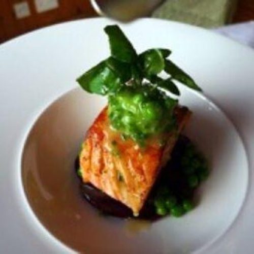 A small piece of smoked salmon sits on a white plate on top of green puree and is garnished with a small green leaf