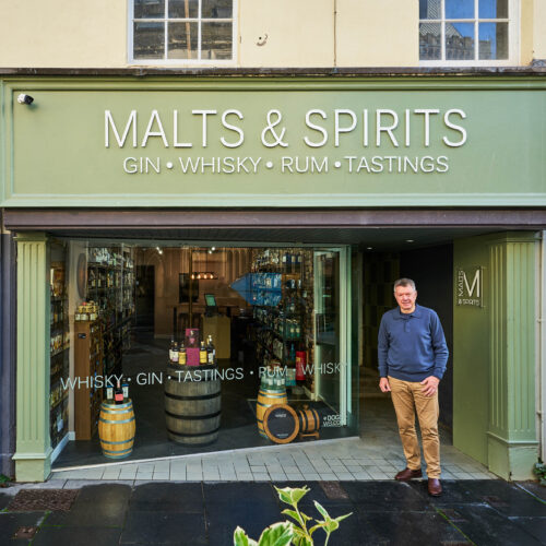 A man wearing a grey jumper stands outside a shop painted light green with a large window displaying bottles of alcohol
