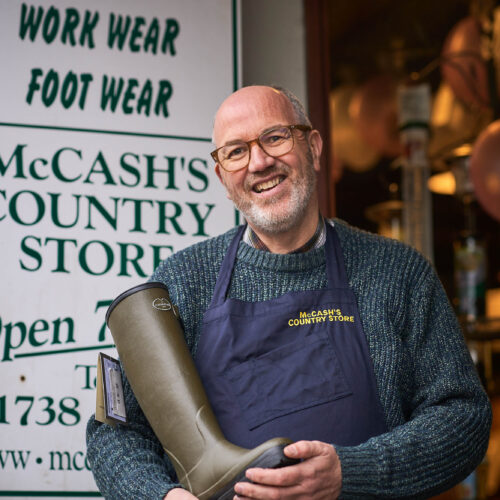 A smiling man wearing a blue apron holds a Wellington boot in front of a white sign with green font outside a shop in Perth