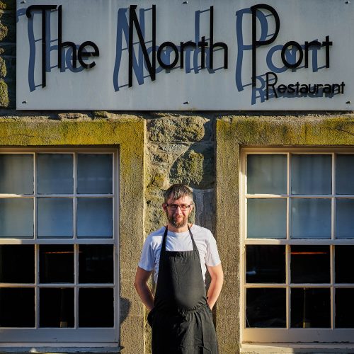 A man wearing a white shirt and black apron stands outside a restaurant with two windows and a large grey sign with a black font