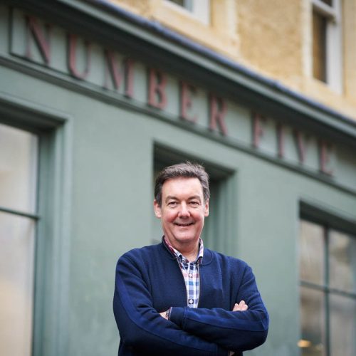 A smiling man wearing a blue jumper and checked shirt stands with his arms crossed outside a grey shop