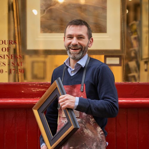 A smiling man wearing a blue jumper under a dirty red apron holds an empty picture frame outside a shop with red trimmings