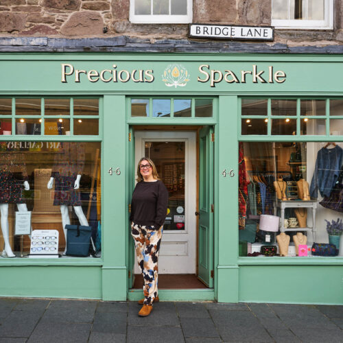 A smiling woman wearing patterned clothing leans against a light green shop with two large windows displaying clothing items