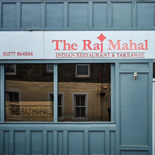 A grey restaurant front with a white sign with red writing above a large window.