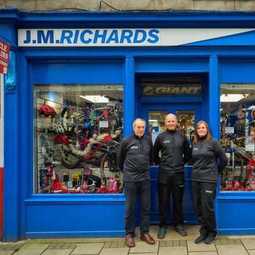 Two men and a woman wearing black stand together outside a blue shop with two windows displaying bikes and cycling supplies