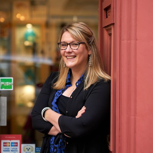 A smiling woman wearing black glasses and a black and blue shirt leans against a red framed shop window