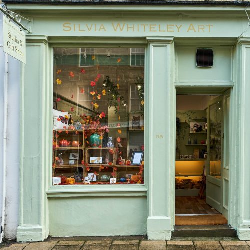A pale green shop with gold lettering reading Silvia Whiteley Art above a large window decorated with autumnal leaves and art
