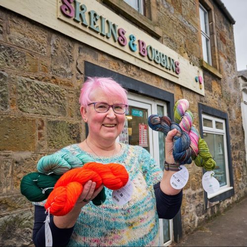 An older woman with pink hair wearing a blue jumper holds colourful yarn outside a shop with a multi-coloured sign