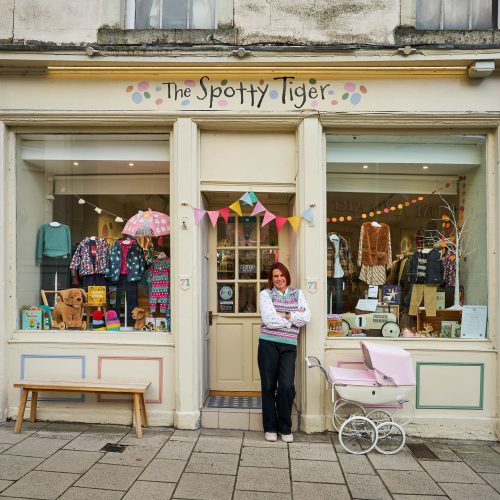 A woman wearing a bright, patterned jumper stands outside a brightly coloured shop displaying children's clothes and toys