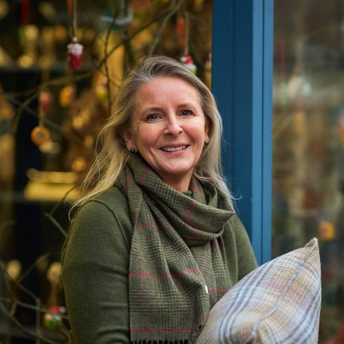 A smiling woman with blonde hair wearing a green plaid scarf holds a white plaid pillow outside a decorated shop window