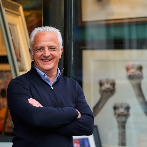 A smiling man with white hair wearing a blue jumper stands with his arms crossed outside a window displaying artwork in Perth