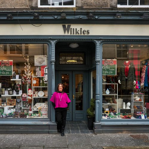 A smiling woman wearing a pink jumper stands outside a shop with two large windows displaying assorted clothing and small gifts