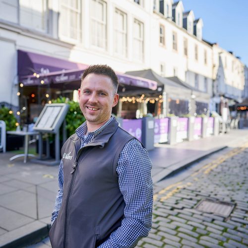 A smiling man wearing a grey shirt and jumper stands outside a cafe with purple signs and a large canopy in Perth