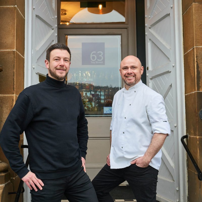 A man wearing a black jumper stands next to a man wearing a chef's uniform outside a the white doorway of a restaurant in Perth