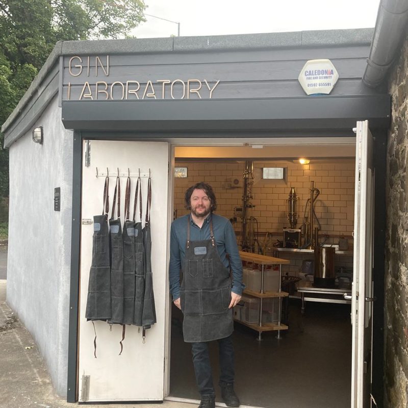 A man wearing a blue shirt and black apron leans on a doorway which opens into a room filled with distillery equipment in Kinross