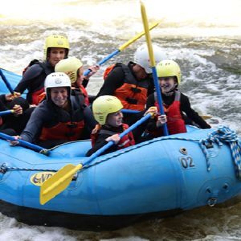 Lots of smiling people holding paddles in a large kayak in fast flowing water.