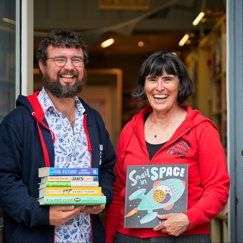 A smiling man wearing a black jacket standing next to a smiling woman wearing a red jacket, both holding books.