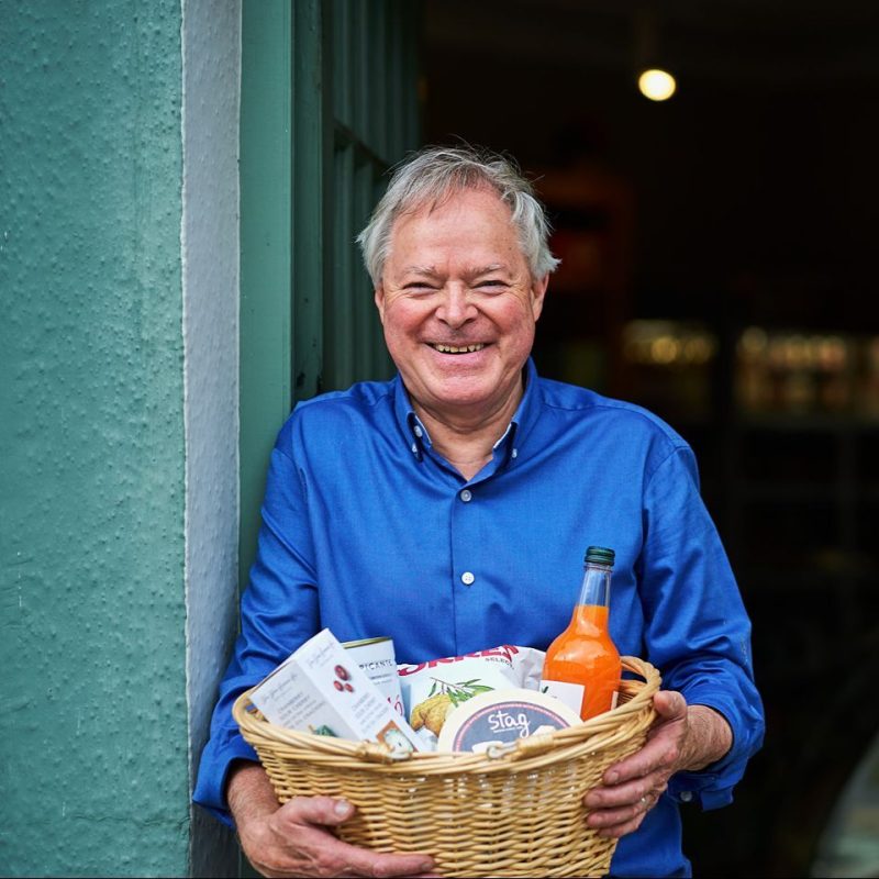 An older man wearing a blue shirt holds a wicker basket filled with several packets of food and a bottle of an orange drink