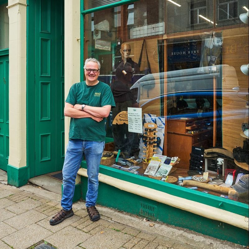 A smiling man with crossed arms wearing a green shirt stands outside a green shop with a large window display of outdoor clothing.