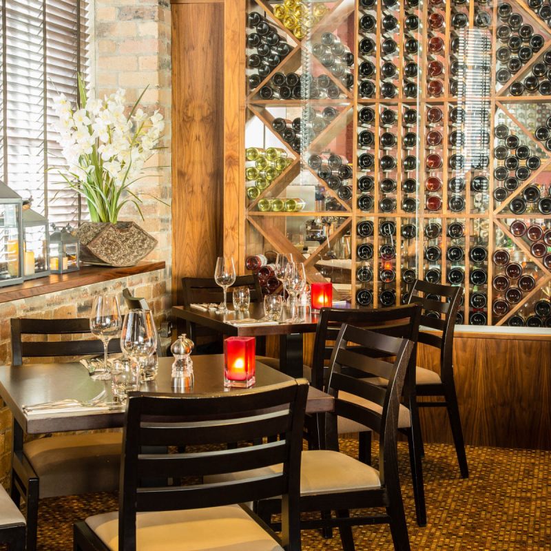 A restaurant in Perth furnished with black dining tables, chairs, wine glasses, and a tall wooden shelf holding several bottles of wine