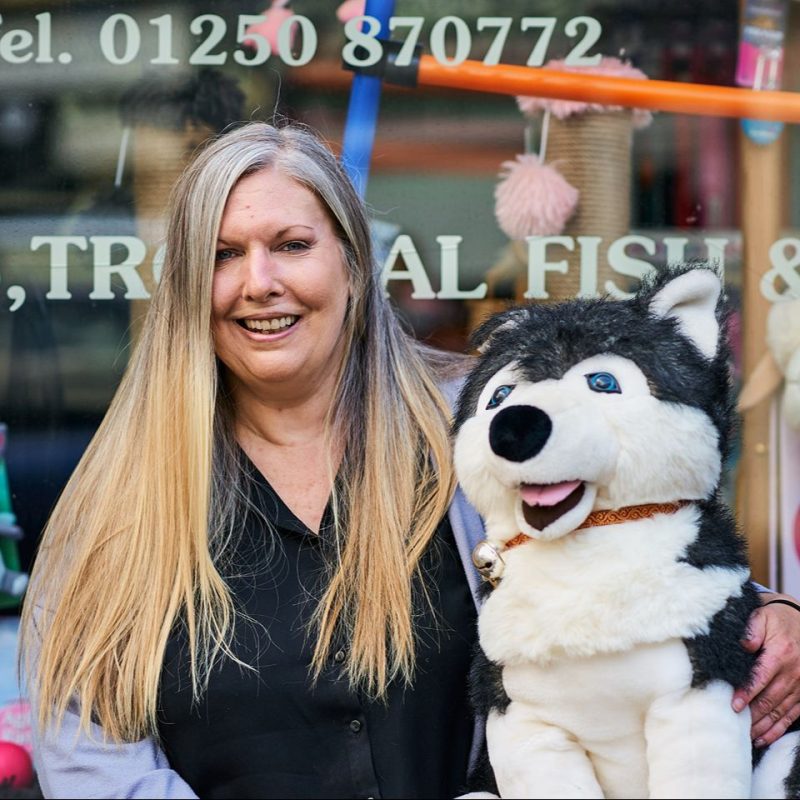 A smiling woman wearing a black shirt holds a large black and white toy dog outside a shop