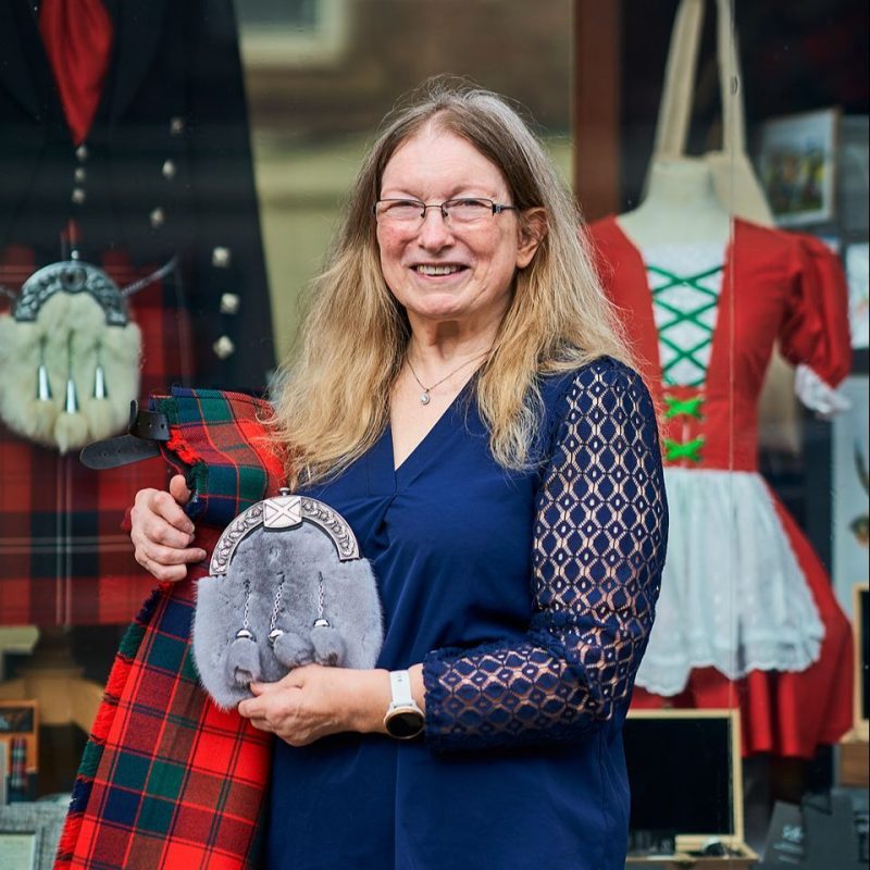 A smiling woman wearing a blue shirt holds a red kilt and grey sporran outside a shop window displaying other Scottish items.