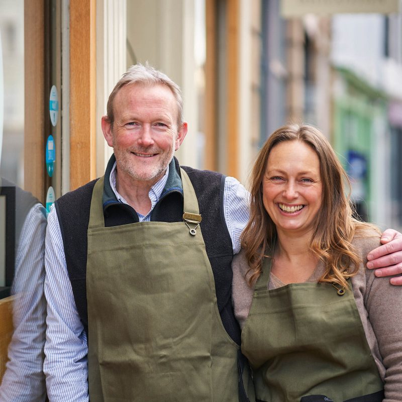 A smiling man rests his hand on a smiling woman's shoulder, both wearing a khaki apron