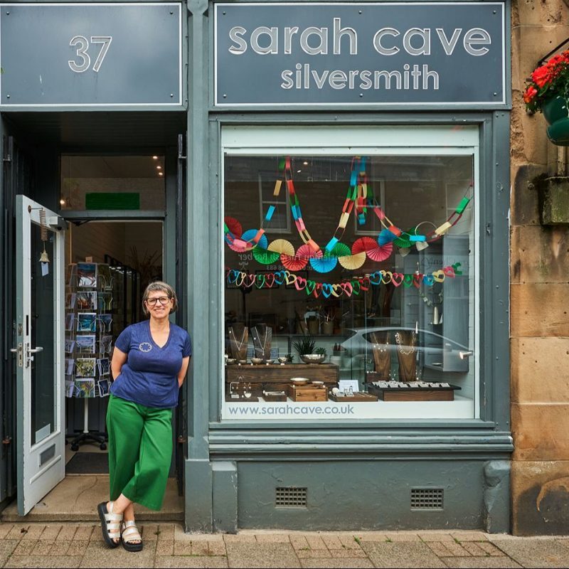 A woman wearing a blue shirt leans next to a large grey window displaying jewellery outside Sarah Cave Silversmith