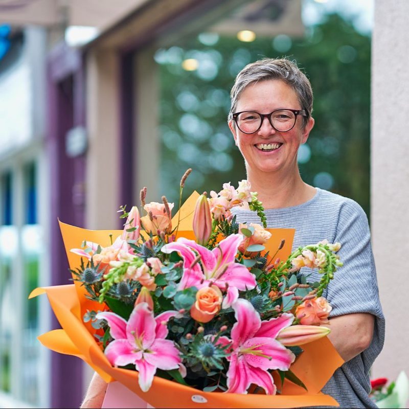 A smiling woman wearing a grey shirt holds a large bouquet of brightly coloured flowers.
