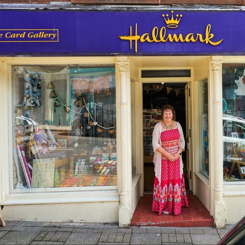A smiling woman in a bright pink dress stands outside The Card Gallery next to a large window display full of craft supplies