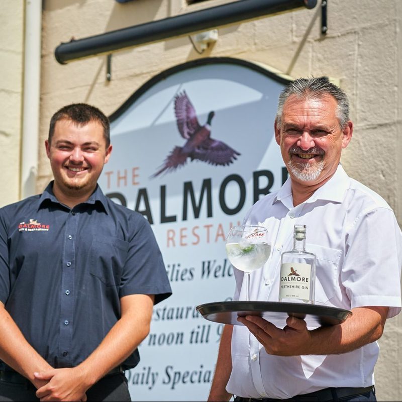 man in white shirt with grey beard holding a tray with gin standing beside a younger man in a blue shirt outside a Blairgowrie hotel