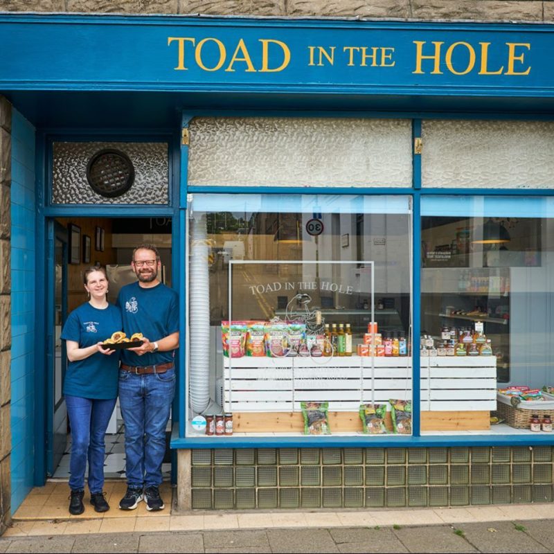 A man and a woman in matching blue shirts stand outside Toad in the Hole next to a large window filled with grocery goods.