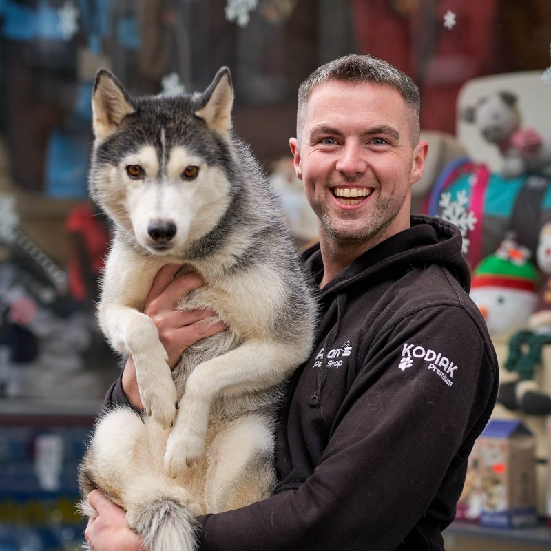 A smiling man wearing a grey jumper holds a large white and grey husky outside a pet shop