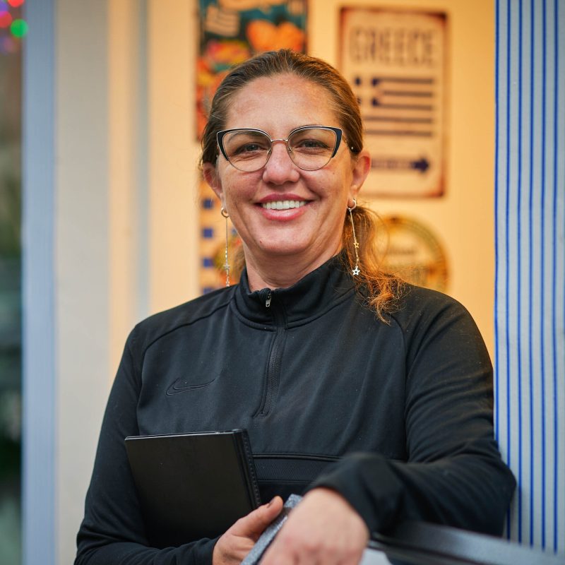 A smiling woman wearing glasses and a black jumper holds a black journal and stands in a restaurant doorway