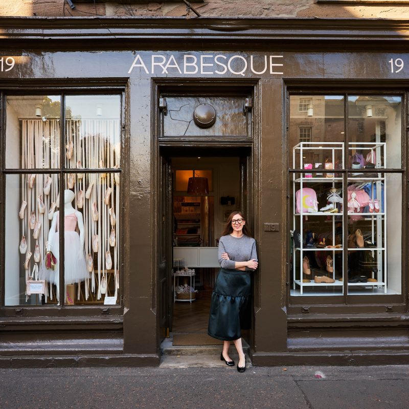 A smiling woman wearing a grey shirt and green skirt leans against a large brown shop window displaying ballet products