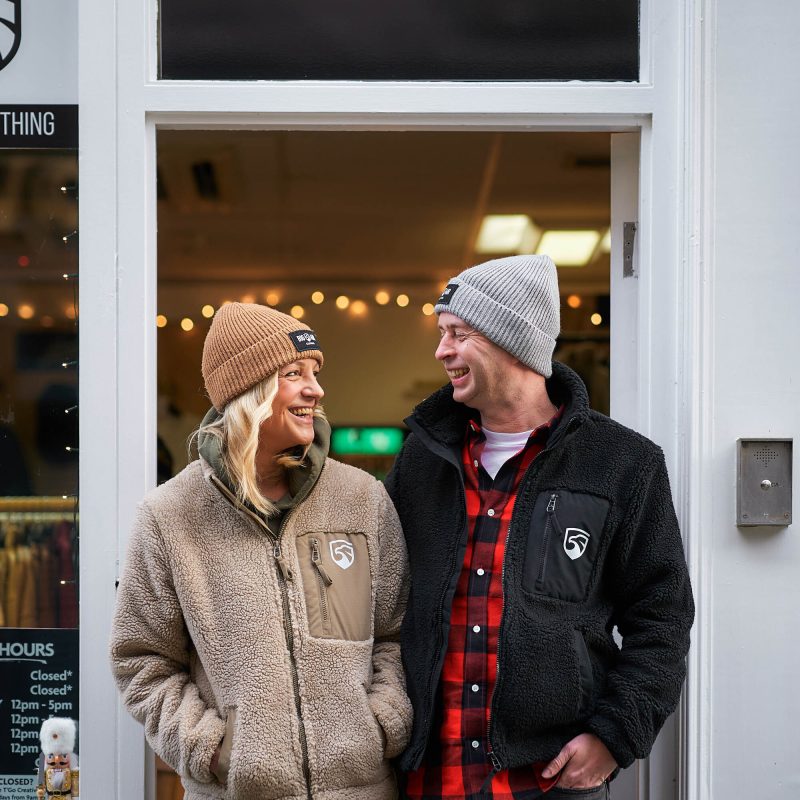 A man and woman wearing hats smile at each other outside a white doorway