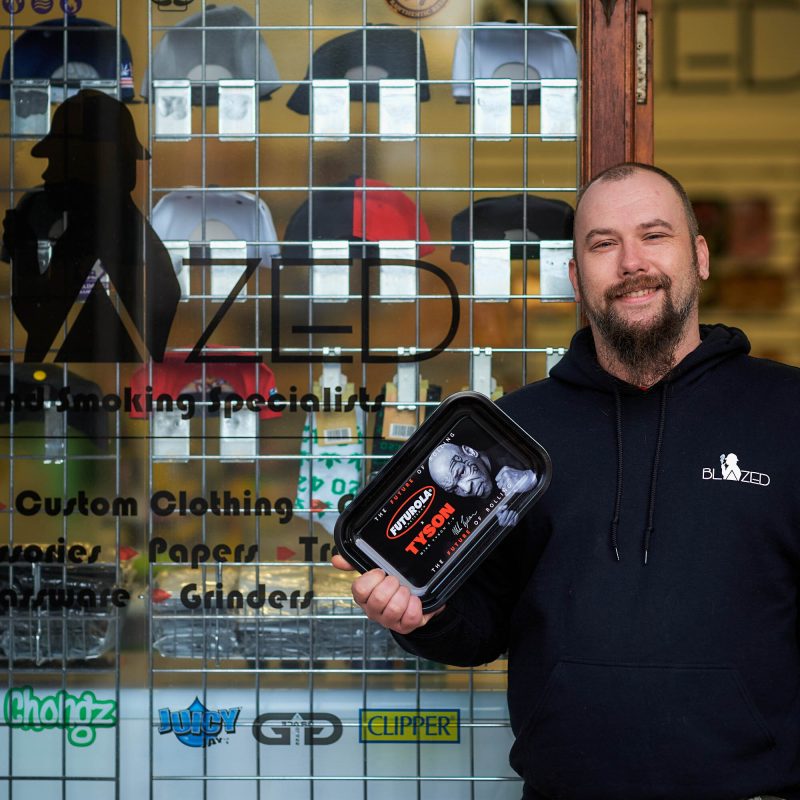 A smiling bearded man wearing a black jumper holds a black tray and stands outside a shop window displaying baseball caps