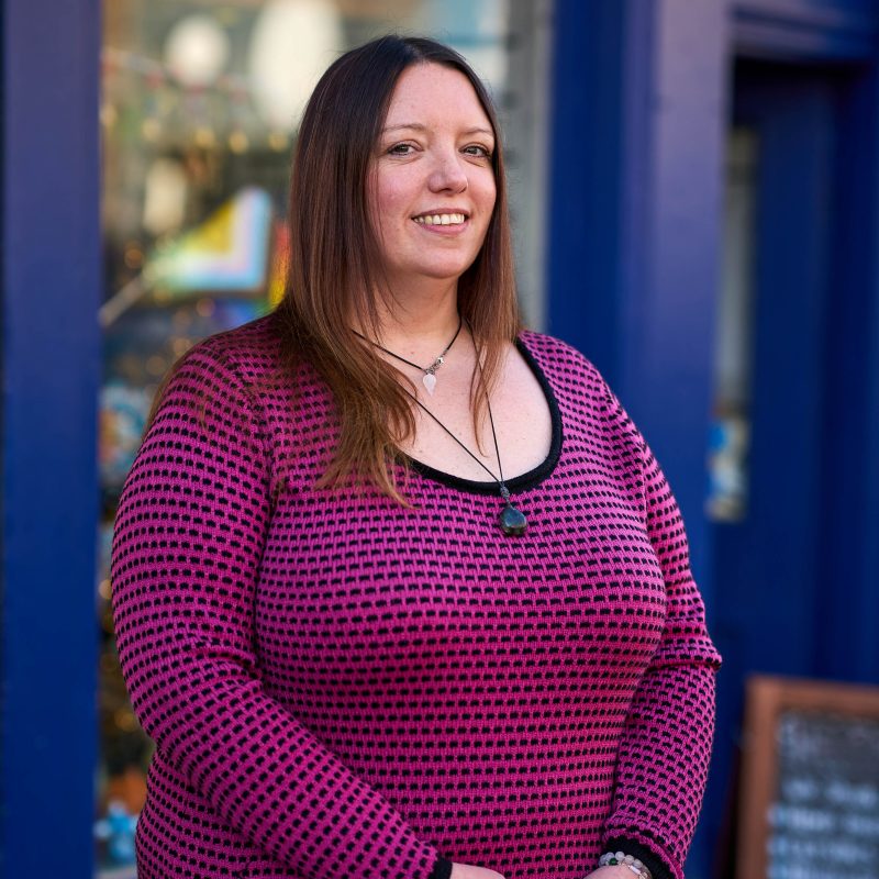 A smiling woman with brown hair wearing a pink and black patterned shirt stands outside a shop painted blue